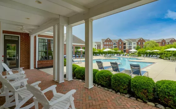 Covered patio with white Adirondack chairs overlooks a large outdoor swimming pool surrounded by lounge chairs and apartment buildings.