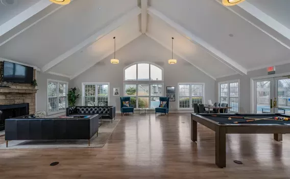 Spacious clubhouse common room with a pool table, dark sofas, blue accent chairs, and large windows under a vaulted ceiling.