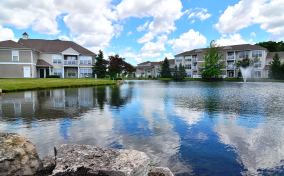 Residential buildings reflected in a tranquil lake under a blue sky with clouds. Rocks in foreground, fountain in distance.