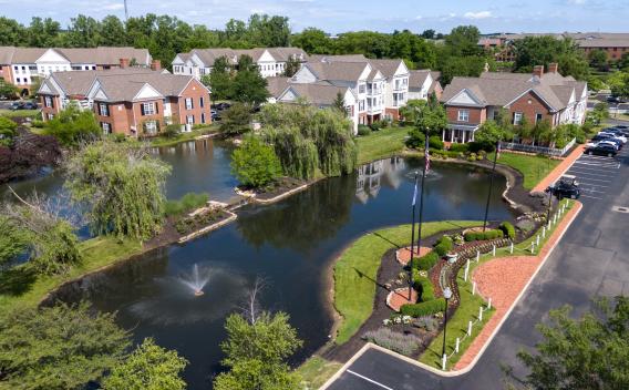 Aerial photograph of a residential community with a large pond, fountains, weeping willow, and brick townhouses under a blue sky.
