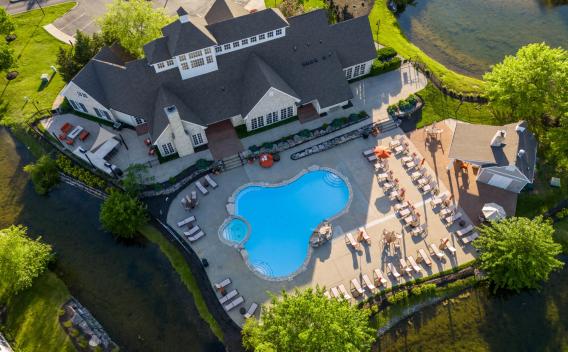 Aerial view of a clubhouse with a freeform swimming pool surrounded by lounge chairs, green lawns, trees, and two ponds.