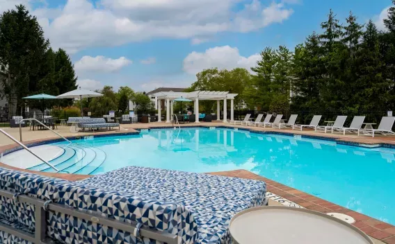 Sunny outdoor swimming pool with blue water, surrounded by lounge chairs and green trees under a partly cloudy sky.