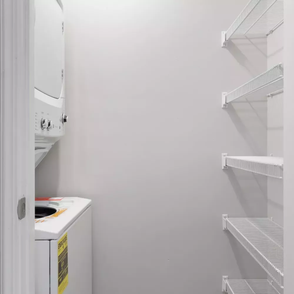 White stacked washer/dryer in a narrow laundry closet with wire shelving and a louvered door.