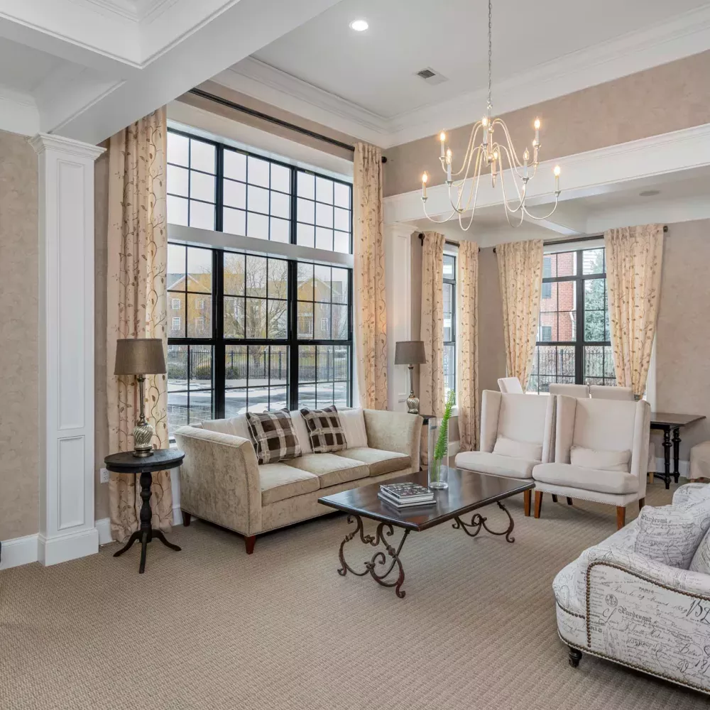 Elegant lounge with a long sofa, armchairs, coffee table, and large windows with patterned curtains. Chandelier overhead.
