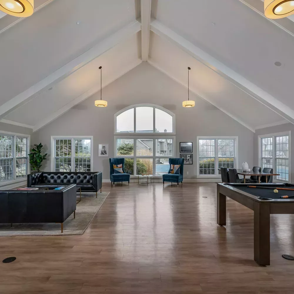 Spacious clubhouse common room with a pool table, dark sofas, blue accent chairs, and large windows under a vaulted ceiling.