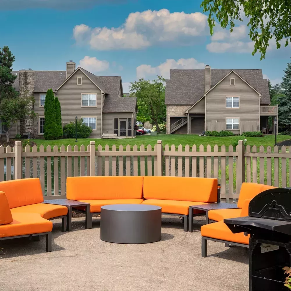Outdoor patio with bright orange seating, a grill, and tables, overlooking a pond with a fountain and residential buildings.