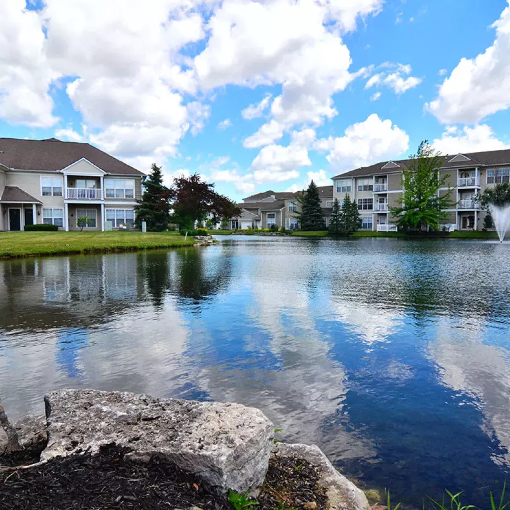 Residential buildings reflected in a tranquil lake under a blue sky with clouds. Rocks in foreground, fountain in distance.
