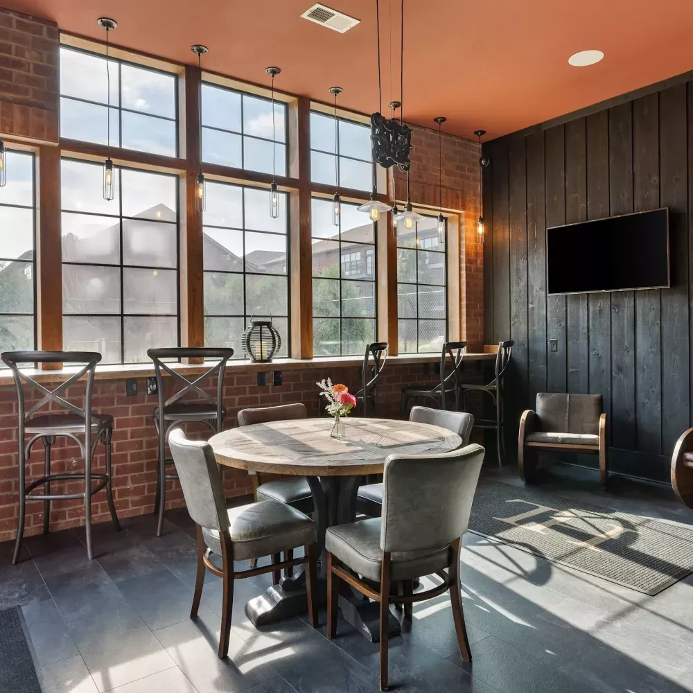 Sunlit modern lounge with large windows, exposed brick, a round table, and a dark wood paneled wall with TV and lounge chairs.