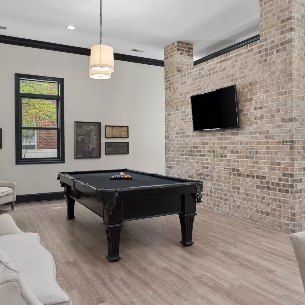 Modern game room featuring a black pool table, light sofa, large window, and a rustic brick wall with a mounted TV.