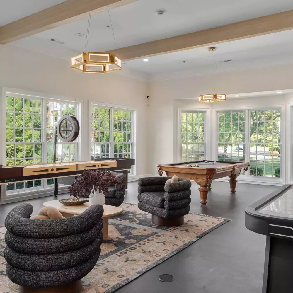 Modern recreation room with pool table, air hockey, and shuffleboard. Plush armchairs, coffee table, and large windows.