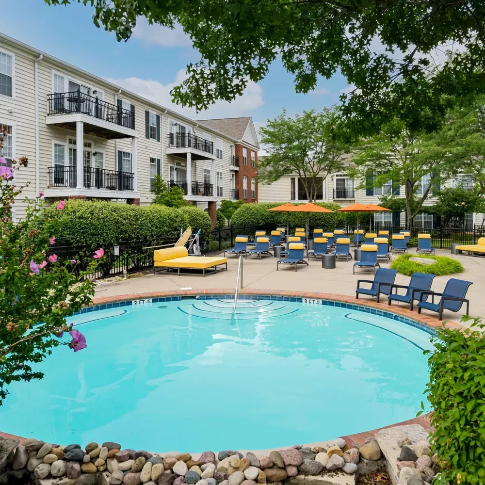 Apartment complex pool with blue water, yellow lounge chairs, blue umbrellas, and a building with balconies.