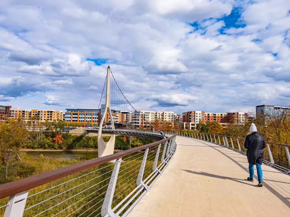 Person walks on a modern suspension bridge over a river, with colorful buildings and autumn trees under a cloudy blue sky.