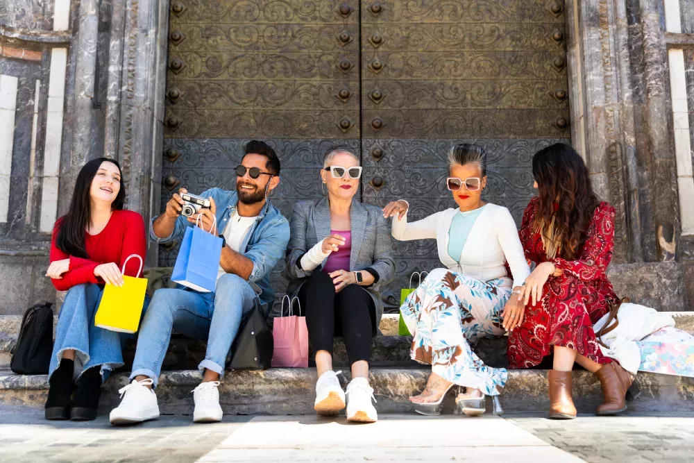 Five diverse friends sit on stone steps after shopping, holding colorful bags. One man takes a photo.