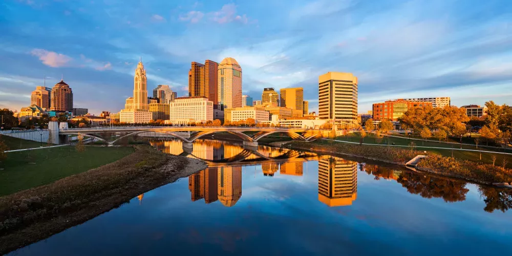 A city skyline at sunset, with tall buildings reflected in a river. A bridge crosses the water, and trees show autumn colors.