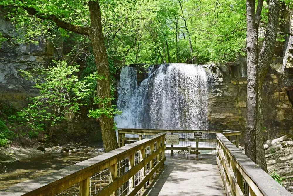 Photograph of a waterfall cascading over rocks, viewed from a wooden platform, surrounded by lush green trees.