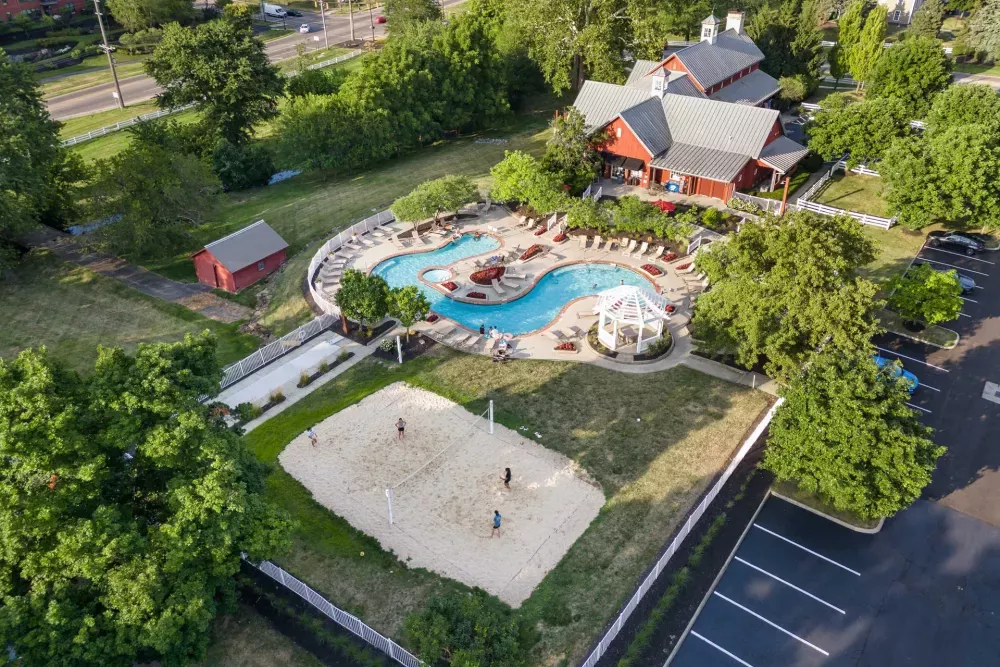 Aerial view of a recreational area with a red clubhouse, blue pool, and sand volleyball court with people playing.